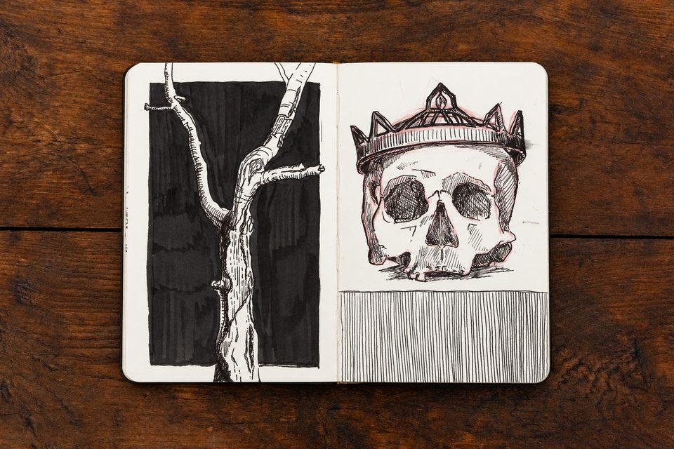 Open sketchbook on a wooden tabletop, with an ink drawing of a tree branch and a crowned skull.